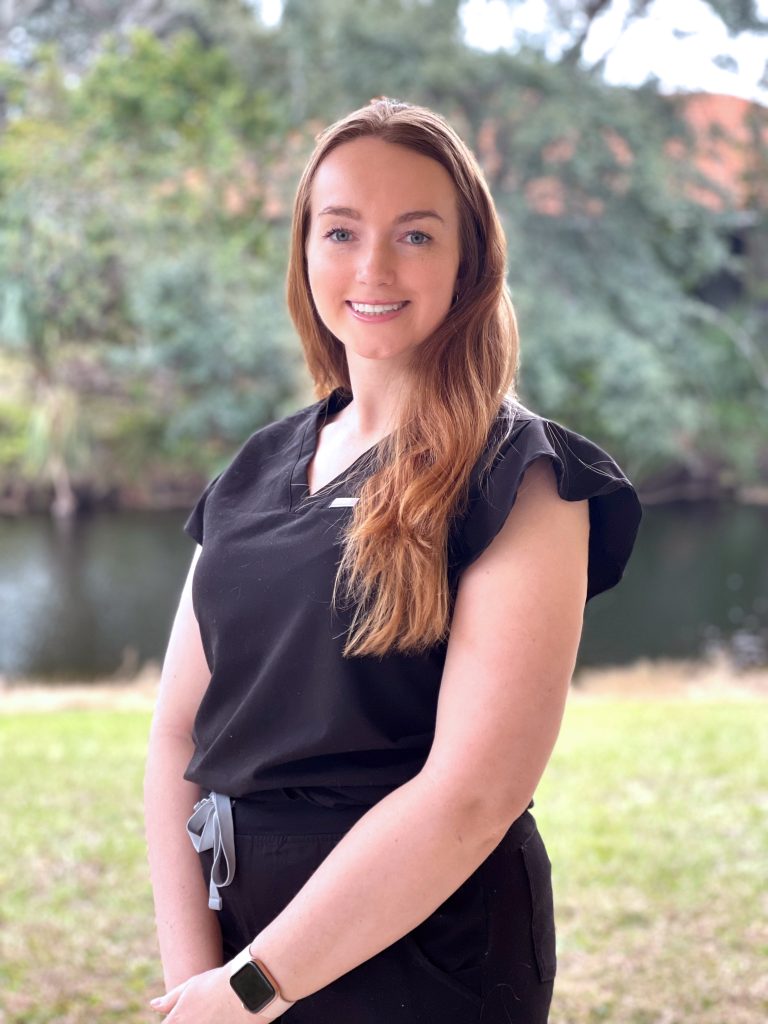 A woman with long hair stands outdoors by a body of water, wearing a black outfit and smiling at the camera.